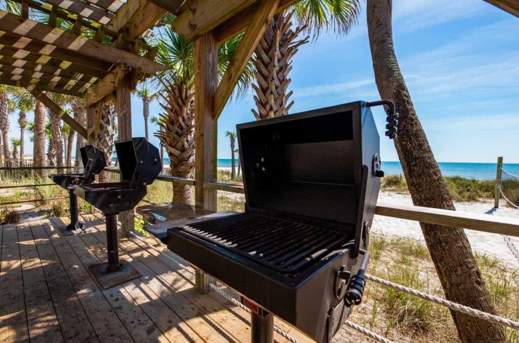 A black barbecue grill on a wooden deck under a pergola with palm trees and ocean view in the background.